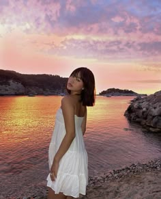 a woman in a white dress standing on the beach at sunset with her eyes closed