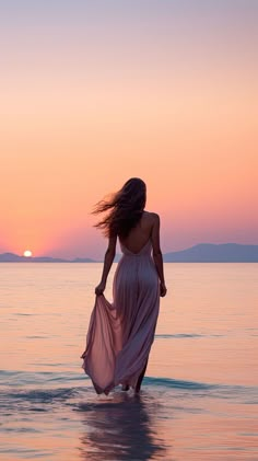 a woman walking in the water at sunset with her dress blowing in the wind,