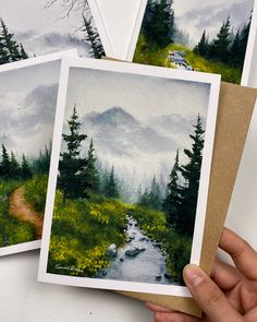 hand holding three cards with watercolor painting of mountains and trees in the background on brown paper