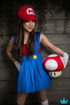 a woman in a blue dress and red hat holding a nintendo mario mushroom stuffed animal