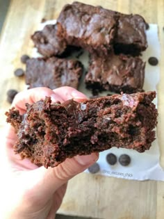 a person holding up a chocolate brownie in front of some other food on a cutting board