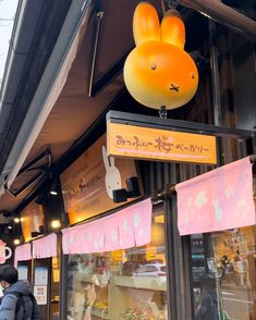 an orange bunny head hanging from the side of a store front with people walking by