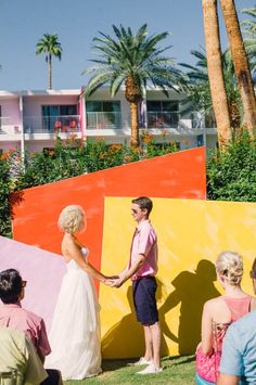 two people standing next to each other in front of colorful walls and palm trees, with one person holding the hand of another persons's hand