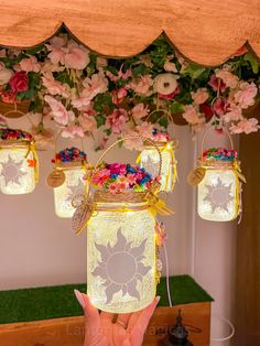 a person holding up a mason jar with flowers hanging from it's sides and lights on the ceiling