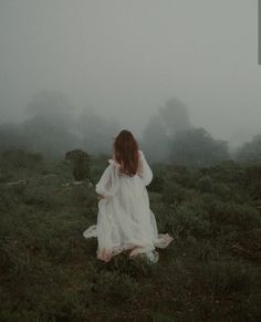 a woman in a white dress walking through a field on a foggy, overcast day