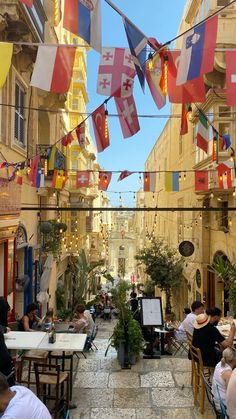 people are sitting at tables in an alleyway with flags hanging from the buildings above