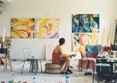 a woman sitting on a chair in front of some paintings and other art supplies around her