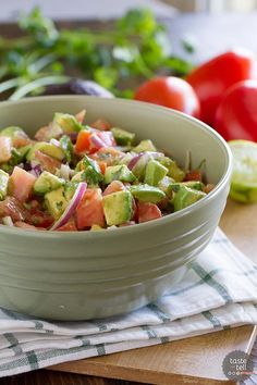 a green bowl filled with lots of food on top of a wooden table next to tomatoes and lettuce