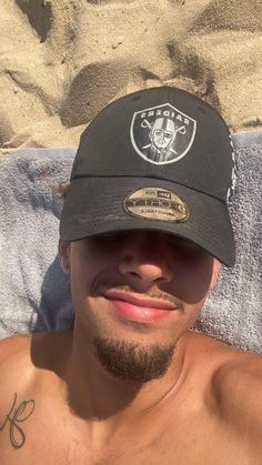 a man laying in the sand wearing a oakland football hat