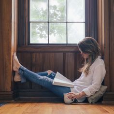 a woman sitting on the floor reading a book with her feet propped up in front of a window