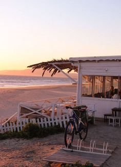 there is a bike parked on the beach next to a small building with people sitting at it