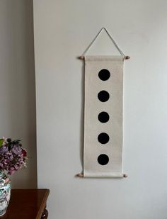 a white wall hanging with three black dots on it next to a vase filled with flowers