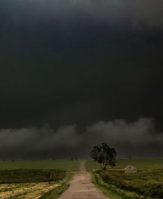 a dirt road leading to a green field under a dark sky with storm clouds in the distance