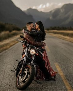 a man and woman sitting on a motorcycle in the middle of an empty road with mountains in the background