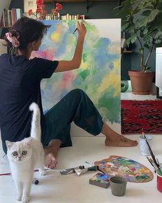 a woman sitting on the floor with a cat and paintbrush in front of her