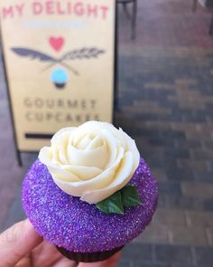 a purple cupcake with white frosting and a rose on top is held in front of a sign that says my delight