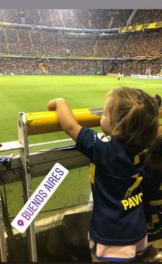 a little boy holding onto a yellow fence at a soccer game in a stadium with people watching from the bleachers