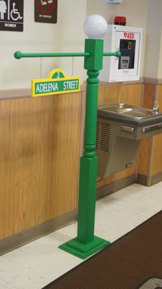 there is a green street sign on the pole in front of the sink and trash can