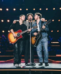 three men standing on stage with guitars and microphones in their hands while one man is holding an acoustic guitar