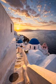 the sun is setting over some white buildings with blue domes on them and water in the background