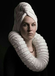 a woman with a white towel wrapped around her head and wearing a black top, standing in front of a dark background