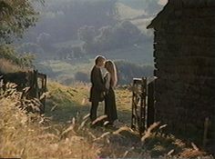 two people standing next to each other in front of a stone wall and gate with grass on the ground