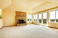 an empty living room with large windows and a fireplace in the center, surrounded by carpeted flooring