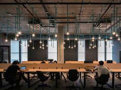 two people sitting at desks in an office setting with computers on the tables and lights hanging from the ceiling