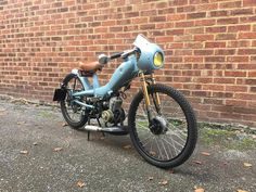 an old blue motorcycle parked in front of a brick wall with leaves on the ground