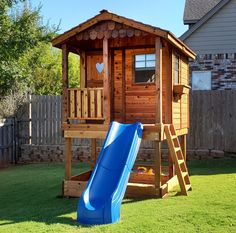 a wooden play house with a blue slide in the grass next to it and a fence