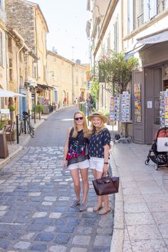 two women standing in the middle of an alleyway with shops on both sides and people walking down the street