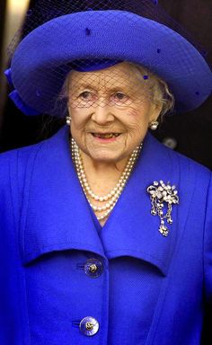 an older woman wearing a blue coat and hat with pearls on the collar, smiling