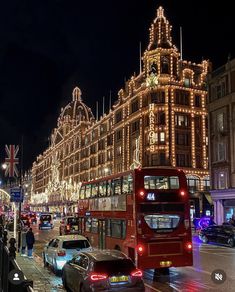 a red double decker bus driving down a street next to tall buildings covered in christmas lights