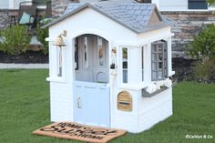 a small white doll house sitting on top of a grass covered field next to a welcome mat