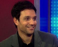 a smiling man in a suit and black shirt on the set of tv talk show