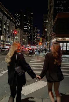 two women holding hands crossing the street in front of some tall buildings at night time