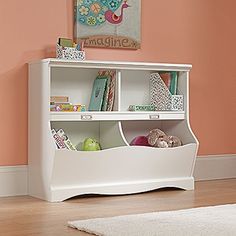 a white book shelf filled with books on top of a hard wood floor next to a pink wall