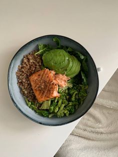 a blue bowl filled with greens and salmon on top of a white tablecloth next to a cup of coffee