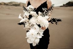 a woman in a black dress holding a bouquet of white flowers and greenery on the beach