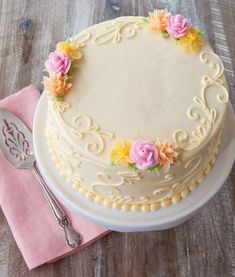 a white cake with yellow and pink flowers sitting on top of a table next to a fork