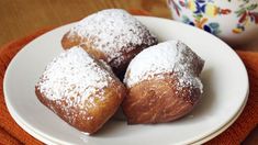 three powdered sugar donuts on a white plate next to a coffee cup and orange towel