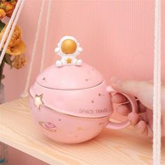 a pink space travel teapot on a wooden shelf with flowers in the back ground