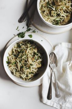 two bowls filled with pasta and vegetables on top of a white tablecloth next to silverware