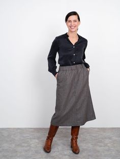 a woman standing in front of a white wall wearing a black shirt and grey skirt