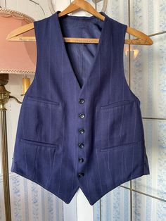 This tailored vest is made from a wool blend in dark royal/navy blue, with widely spaced light blue and beige pinstripes. It has the original buttons in the front and four front pockets, and is fully lined. There are no labels. The measurements, taken with the vest lying flat. are: shoulder to shoulder, 11 inches; armpit to armpit, 19 inches; length, 24 inches in front and 20 inches in back; bottom edge, 17 inches. In very good condition. Sleeveless Tailoring Vest With Welt Pockets, Wool Vest With Pockets For Tailoring, Elegant Wool Vest With Pockets, Classic Blue Three-piece Suit With Pockets, Classic Blue Three-piece Suit, Navy Sleeveless Vest With Pockets, Classic Navy Sleeveless Vest, Fitted Wool Vest With Welt Pockets, Fitted Navy Sleeveless Vest