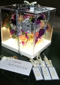 a clear box with flowers inside and tags attached to the sides, on top of a glass table