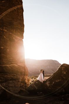 a bride and groom standing in front of a rock formation with the sun behind them