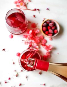 two wine glasses filled with red liquid next to some pink flowers and petals on a white surface