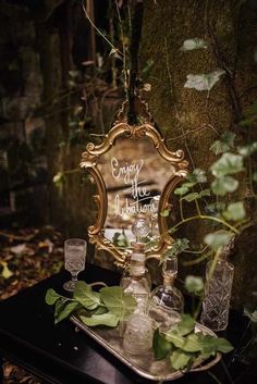 a mirror and some glasses sitting on a table in the woods with ivy growing around it