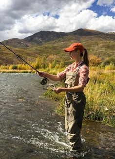 a woman standing in the water while holding a fishing rod and wearing an orange hat